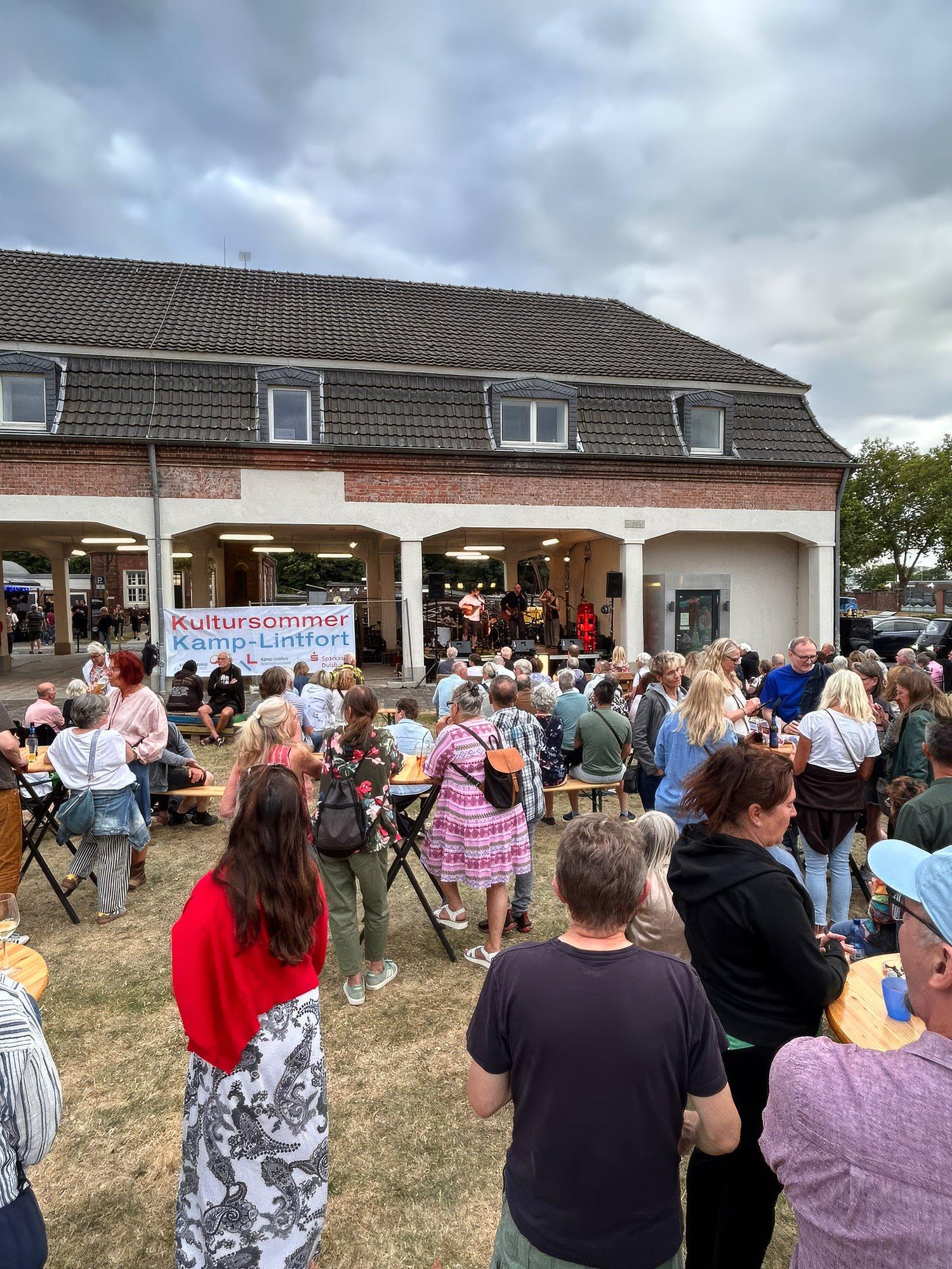 Publikum sitzt und steht bei einem Konzert unter dem Banner Kultursommer Kamp-Lintfort vor dem Schirrhof