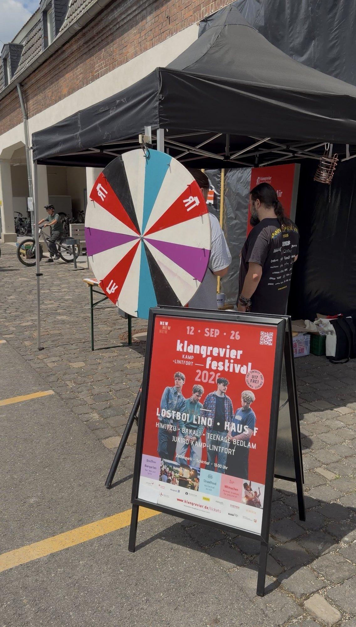 Rot-weißes Glücksrad unter einem Zelt, daneben eine Person und ein Aufsteller des klangrevier music festivals