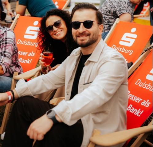 Ein Paar mit Sonnenbrillen sitzt entspannt in orangefarbenen Liegestühlen und stößt mit einem Getränk an
