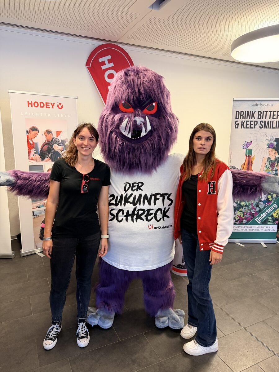 Zwei Mitarbeiterinnen am Messestand von HODEY mit dem Maskottchen DER ZUKUNFTSSCHRECK zwischen sich
