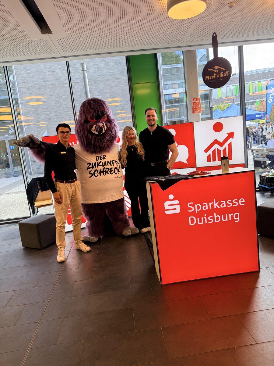 Das Team der Sparkasse Duisburg an seinem roten Messecounter gemeinsam mit dem Maskottchen DER ZUKUNFTSSCHRECK