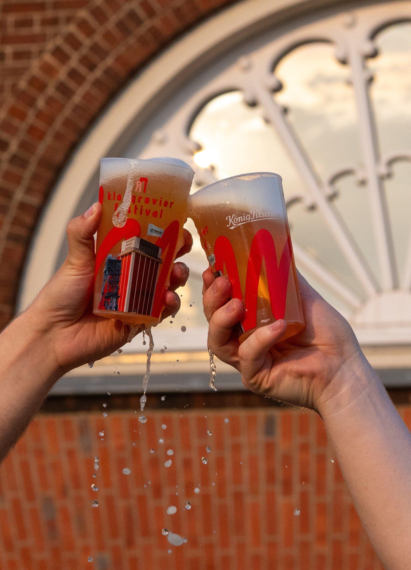 Zwei Becher mit klangrevier-festival-Aufdruck stoßen vor einem Backsteinbogen des Schirrhofs an
