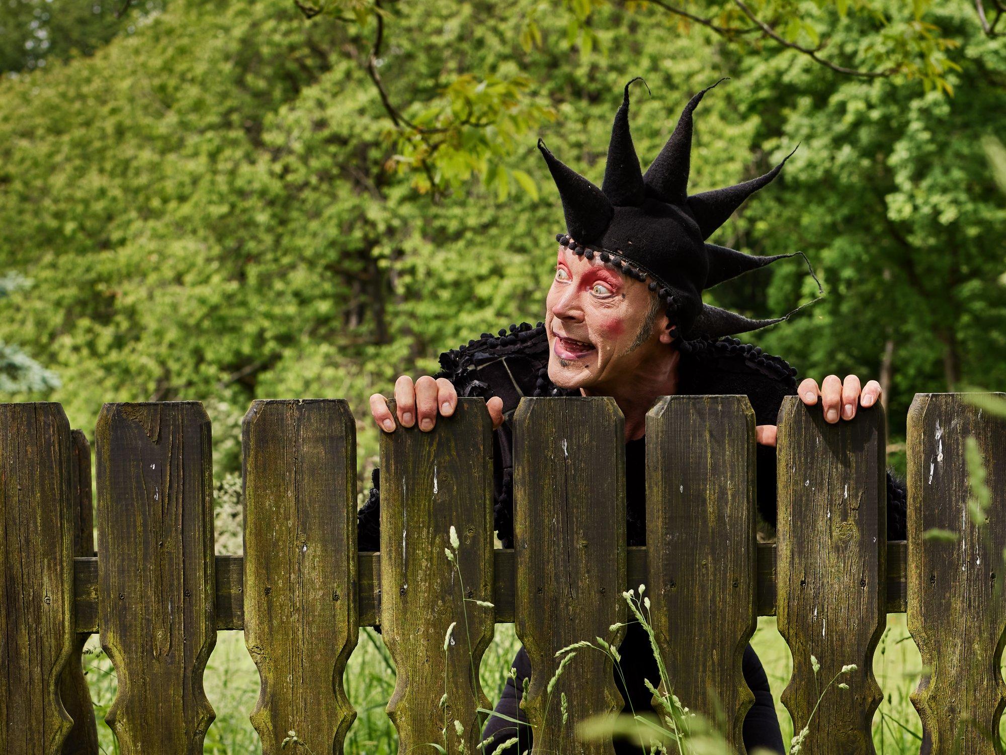 Schauspieler in der Rolle des Till Eulenspiegel mit schwarzer Narrenkappe blickt über einen Holzzaun