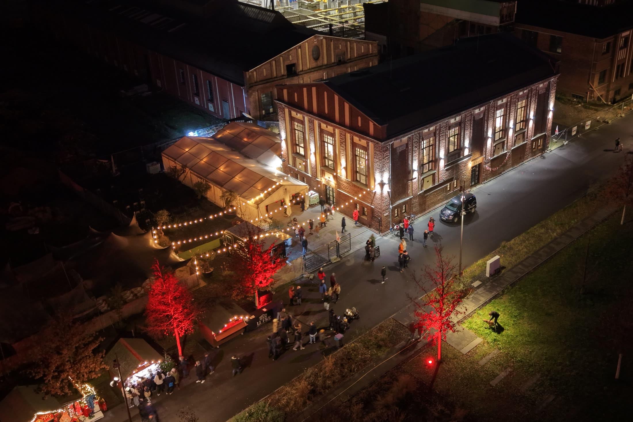 Luftaufnahme des Weihnachtsmarkts im Zechenpark bei Nacht: beleuchtete Hütten, rot angestrahlte Bäume und eine Lichterkette entlang der historischen Zechengebäude
